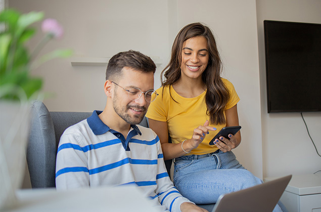 Man sitting in a chair using a laptop with his wife sitting on the arm on her phone, looking at the laptop over his shoulder.